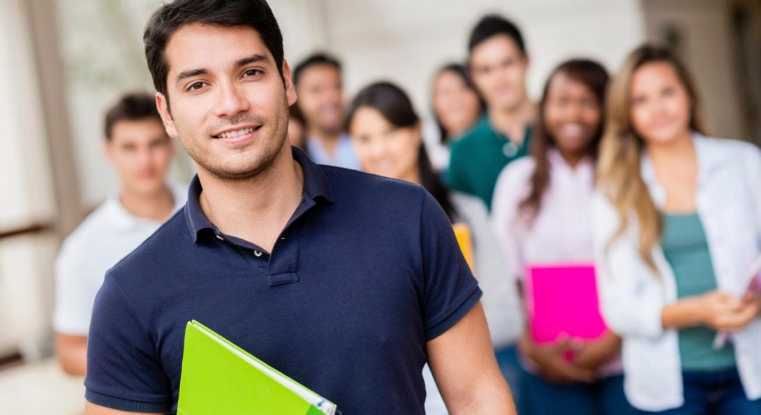 Young man, holding a green notebook smiling with other students in the background